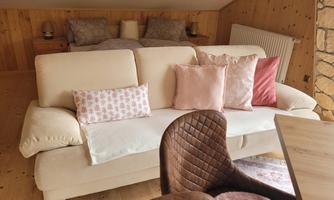 A cozy living room with a bright sofa and decorative pillows. In the background, a bed and wood paneling are visible.