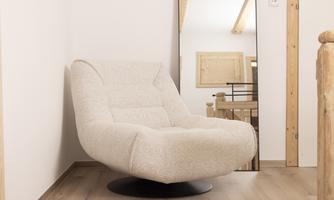A modern armchair in light fabric stands in a cozy corner of a room. In the background, a large mirror and wooden elements are visible.
