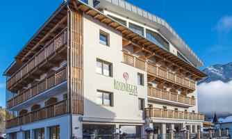 A modern hotel building with wooden cladding and a balcony. The clear sky and the surrounding mountains create an inviting atmosphere.