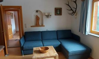 A cozy living room with a blue sofa and a wooden table. The walls are adorned with decorative elements and there is plenty of natural light coming through the windows.