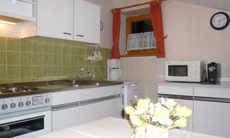 A modern kitchen with white cabinets and green tiles. A flower vase is on the countertop and in the background, there is a window and a clock visible.
