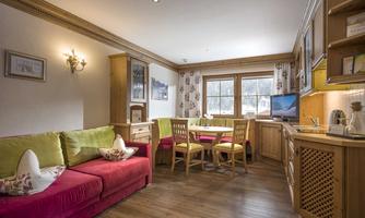 A cozy living space with a red sofa, a table, and wooden chairs. The kitchen is well-equipped and there are large windows that let in plenty of light.