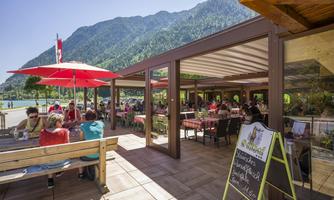 An inviting restaurant by the lake, surrounded by mountains. Many guests are sitting under red sun umbrellas, enjoying the sunshine.
