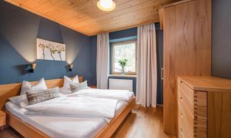 A cozy bedroom with two beds and wooden furniture. The walls are painted in a dark shade, and a window lets in natural light.
