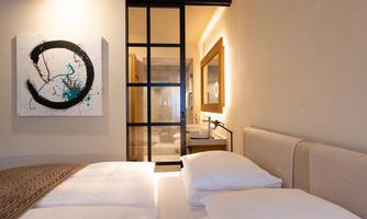 A modern bedroom with two beds and minimalist decor. In the background, a piece of art and a bathroom area are visible.