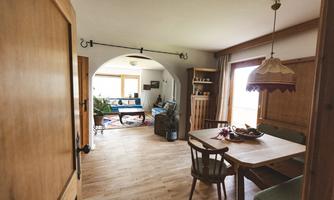 A cozy living space with wooden furniture and plenty of natural light. The room includes a dining area and a seating corner with a sofa.
