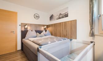 A cozy bedroom with a double bed and a baby cot. The walls are painted in neutral colors and there is wooden paneling behind the bed.