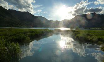 A calm lake surrounded by mountains and green grass. The sun is shining brightly and reflecting on the water.