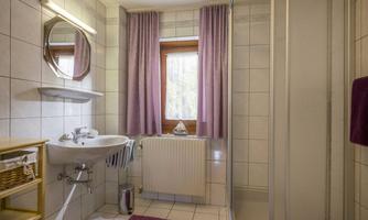 A modern bathroom with white tiles and a large window. There is a shower, a sink, and a small shelf for storage.