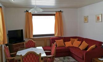 A cozy living space with a red sofa and orange cushions. There is a table with chairs and large windows with yellow curtains.