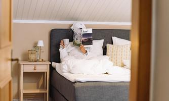 A cozy bedroom with a large bed. A person is relaxed reading a magazine and holding a cup in their hand.