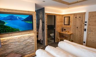 A modern room with wooden walls and a large television. There is a bed, a table, and access to a bathroom.