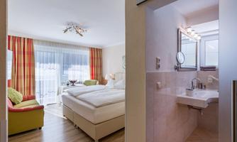A modern room with two beds, a cozy armchair, and large windows. The adjoining bathroom is bright and functionally designed.