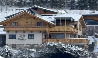 A wooden house in alpine style, surrounded by snow. The terrace is equipped with wooden railings and the surroundings consist of a wintry landscape.