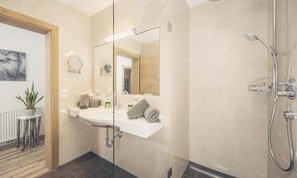 A modern bathroom with a glass shower and bright, minimalist design. There is a plant and towels on the shelf.