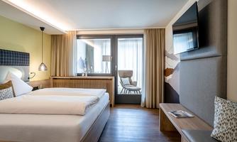 A modern hotel room with a double bed, a television, and a large window. There is a cozy seating area with a chair and plenty of natural light.