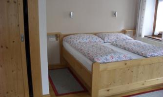 A cozy bedroom with a wooden double bed and colorful bedspreads. The room has wood-paneled walls and a window that lets in daylight.