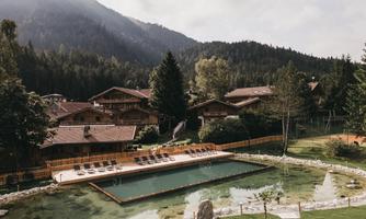 A picturesque mountain landscape with a small swimming pond and cozy wooden chalets. The surroundings are surrounded by trees and mountains, creating a relaxed atmosphere.