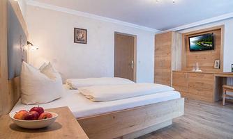 A bright and modern bedroom with a double bed and wooden elements. A bowl of apples sits on the nightstand, and there is a small seating area with a television.