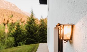 An outdoor area of a house with a lamp that emits warm light. In the background, green trees and mountains can be seen.