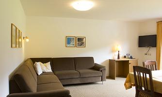 A cozy living room with a beige sofa and a table. Pictures hang on the walls and a television is mounted on the wall.