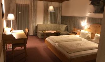 A cozy hotel room with a double bed, a sofa, and a desk. The walls are light, and the room has pleasant lighting.