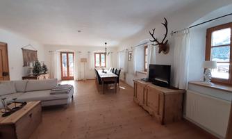 A spacious living room with wooden flooring and large windows. There is a cozy sofa, a dining table, and rustic decorative elements.