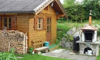 A cozy wooden house with a stone grill next to it. In the foreground lies a small pile of wood and a green meadow.
