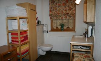 A small bathroom with wooden shelves and colorful towels. There is a window with a patterned curtain and a table with a chair.