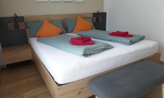 A modern bedroom with a double bed and colorful pillows. On the dresser, there is a towel and the wall is decorated with small pictures.