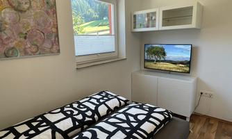 A modern bedroom with two single beds and colorful blankets. A television and a closet are mounted on the wall, and a window allows light from outside.
