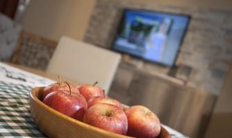 A bowl of fresh, red apples is on a table. In the background, a television is visible.