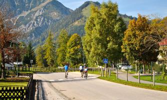 A quiet street with cyclists and many trees. In the background, you can see the mountains under a blue sky.