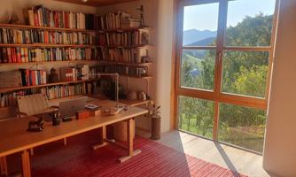 A bright study with a wooden bookshelf full of books. The large window offers a view of nature and the mountains.