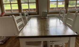 A bright dining room with a wooden table and white chairs. Large windows offer a view of the green landscape and the mountains.