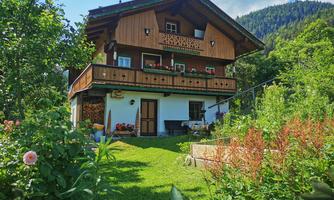 A typical alpine house with wooden cladding and a balcony. The garden is green and blooming, surrounded by mountains.