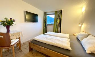 A cozy bedroom with two single beds and a small table. A television and a window with a view of the mountains are visible.