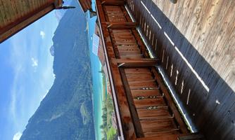A wooden terrace overlooking a beautiful lake and majestic mountains. The landscape is green and sunny, ideal for relaxing.