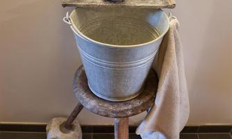 A bucket on a wooden frame with two faucets above it. Next to the bucket hangs a towel.