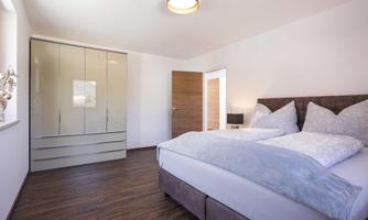A modern bedroom with a large bed and cozy bedding. In the background, there is an elegant wardrobe and a window that lets in a lot of light.