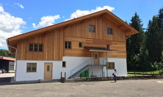 A modern wooden house with a large roof and clear lines. In the foreground, there is a person standing in front of the entrance steps.