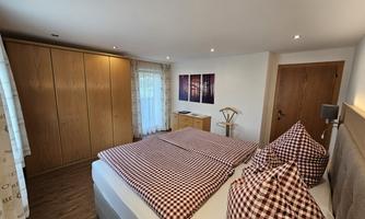 A cozy bedroom with a large bed and checked cushions. There are wooden cabinets and a window with curtains that let in a lot of light.
