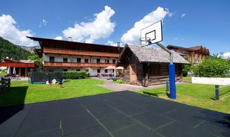 A beautiful outdoor area with a basketball hoop and a green lawn. In the background, rustic buildings and a trampoline can be seen.