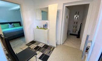 A comfortable entrance area with a chair, a mirror, and a small shelf. In the background, there is a bright bedroom with a bed and colorful cushions.