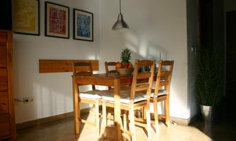 A bright dining area with a wooden table and chairs. A fruit bowl and a plant are on the table in the corner.