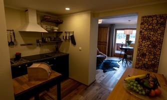 A modern kitchen with wooden shelves and high-quality appliances. In the background, there is a cozy living area with a table and chairs.