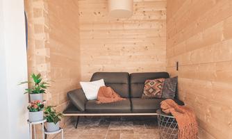 A cozy room with wooden walls and a modern black sofa. There are cushions and a blanket on it, next to it is a plant and a wicker chair.