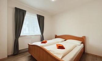 A simple bedroom with a double bed and two orange towels. A window with curtains lets in a lot of light.