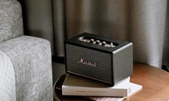 A stylish speaker stands on a table next to a gray couch. The room has a cozy atmosphere with dark curtains.