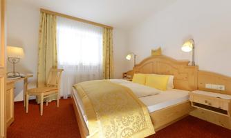 A modern bedroom with a large bed, yellow pillows, and wooden paneling. The room is bright and friendly, with a desk and a comfortable chair.